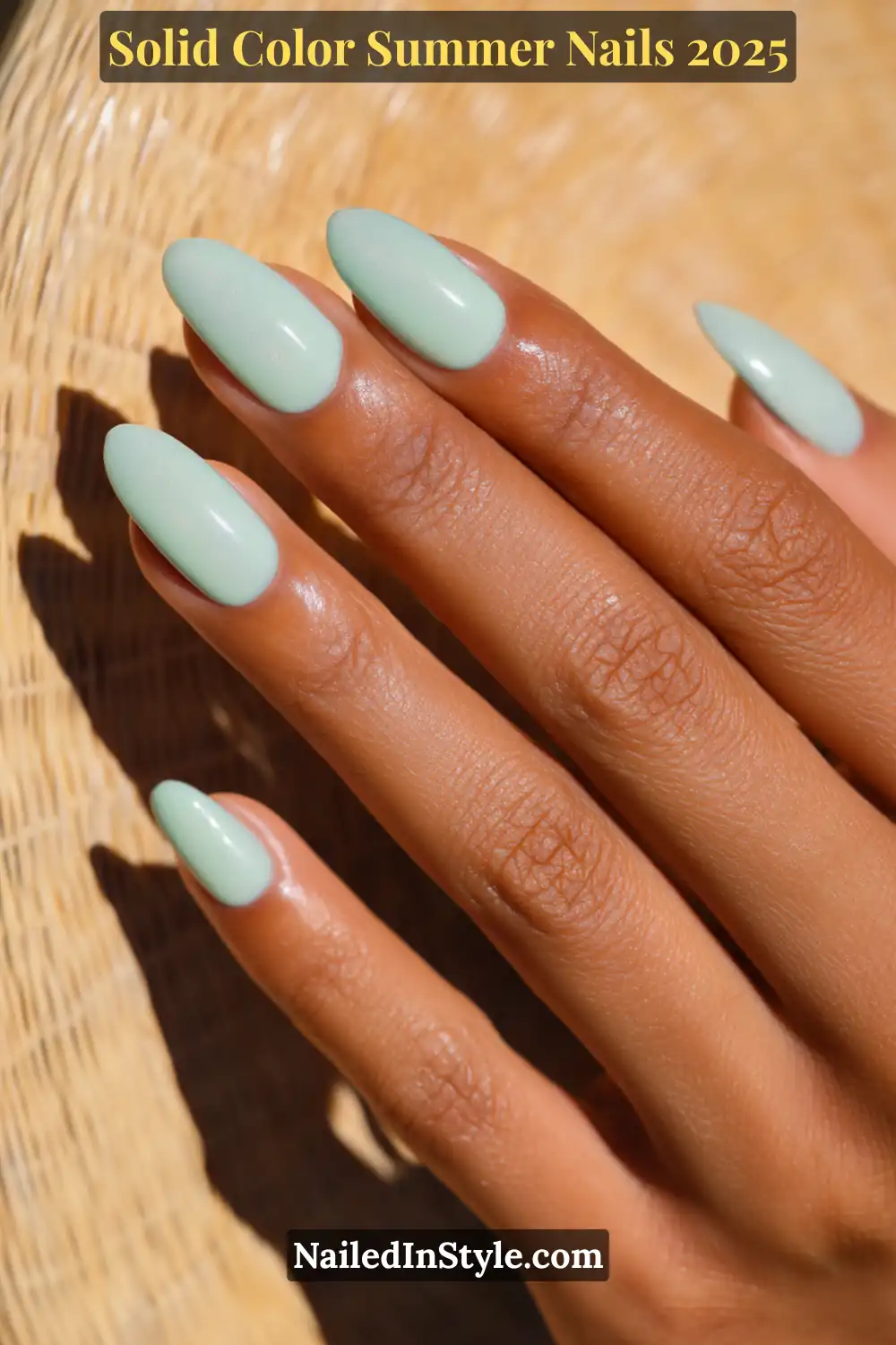 A hand with almond-shaped nails painted in a soft baby mint green with a subtle shimmer finish, shown in sunlight against a woven background.