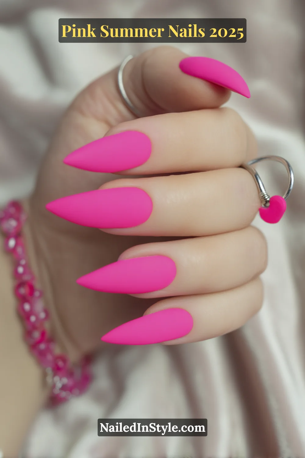 Close-up of a hand with long stiletto nails painted in vibrant matte Barbie pink, accessorized with silver rings and a pink bracelet.