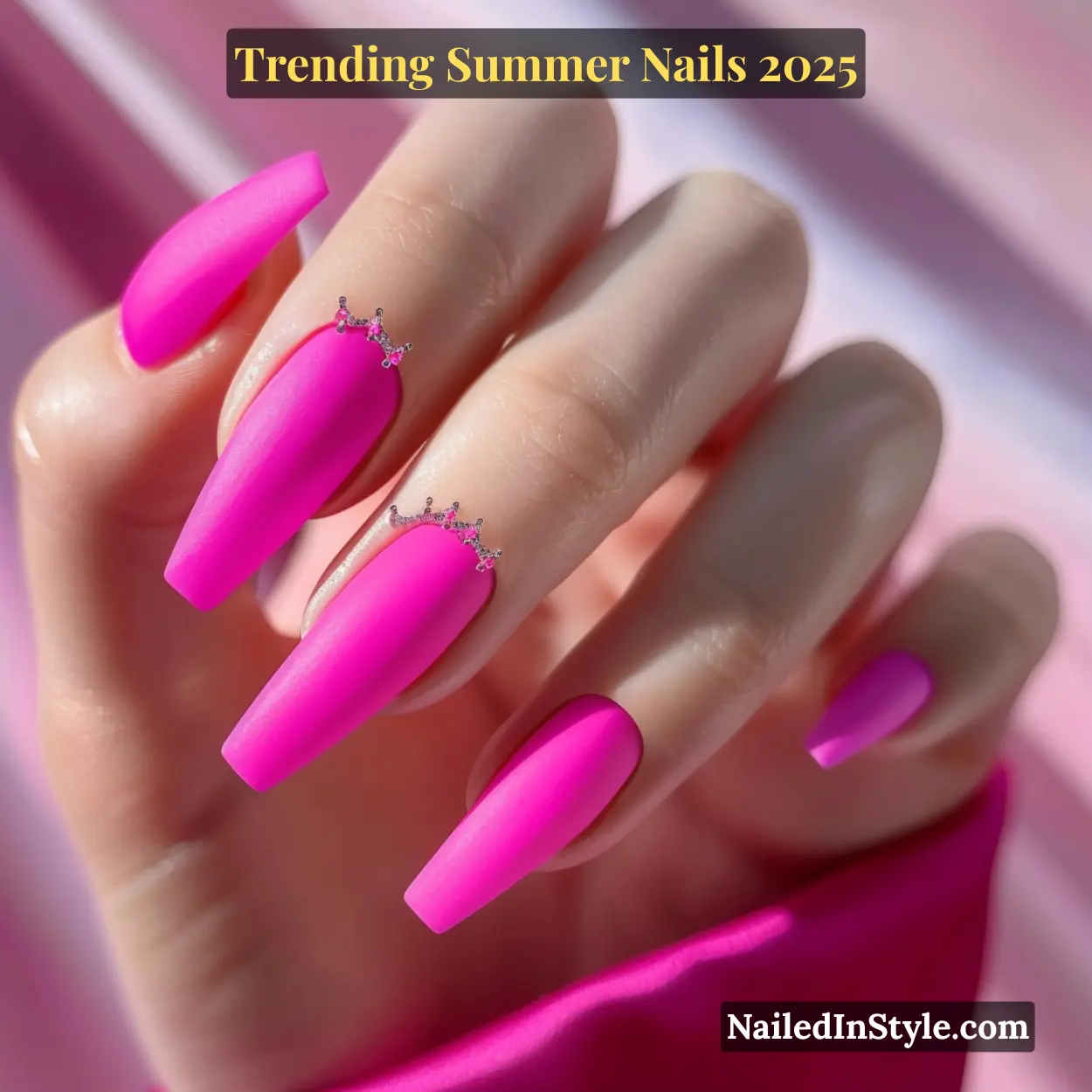 Close-up of a fair-toned hand with long matte hot pink coffin nails, two featuring tiny rhinestone crown embellishments against a soft pink background.