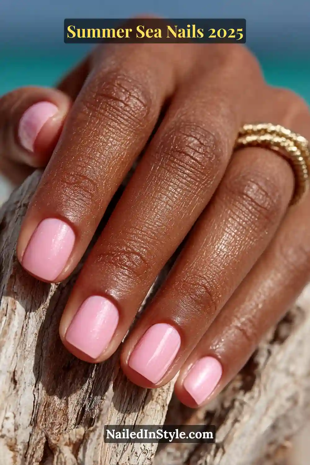 Close-up of natural short nails painted in sheer shell pink BIAB polish, styled with gold rings and beachy background