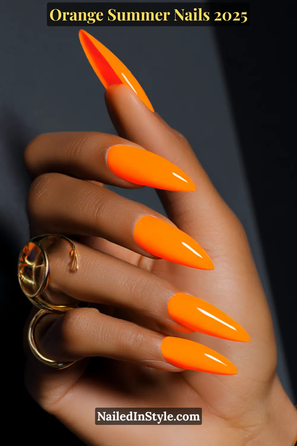 Close-up of neon orange stiletto nails on medium-dark skin tone with a gold circular statement ring against a dramatic dark background.