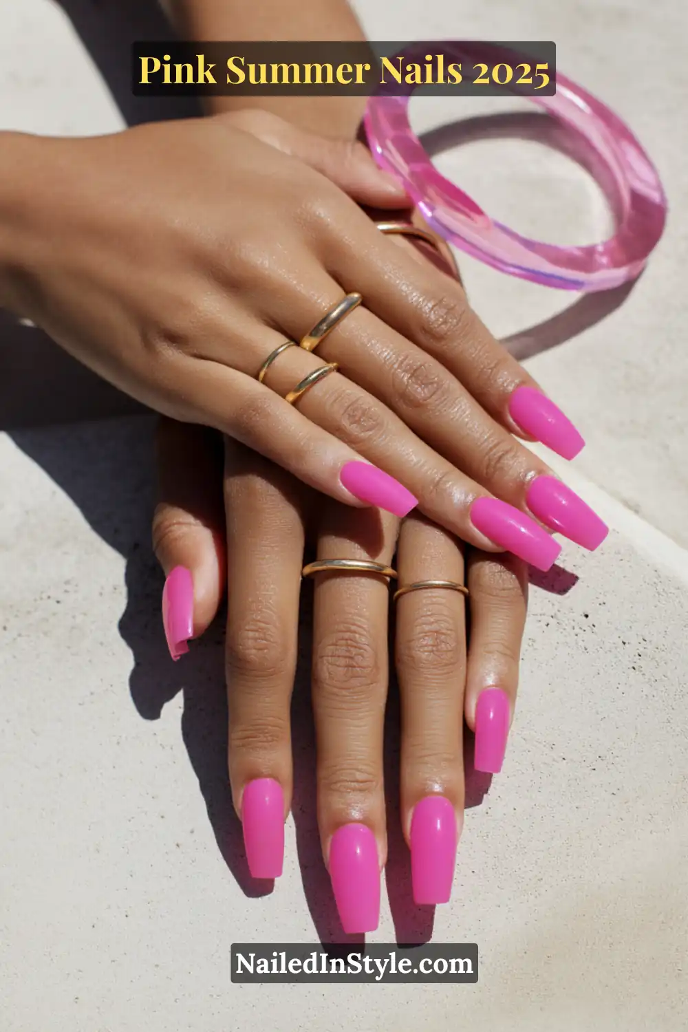 Close-up of hands with long coffin-shaped neon pink nails, styled with gold rings and a translucent pink bracelet for a bold summer 2025 look.