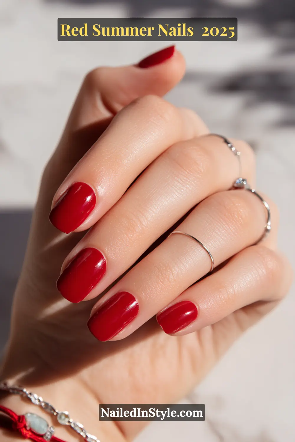 Red Summer Nails 2026: Bold, Fiery & Classic Warm-Weather Looks 13 Close-up of glossy candy apple red squoval nails on fair skin with silver rings and red string bracelet