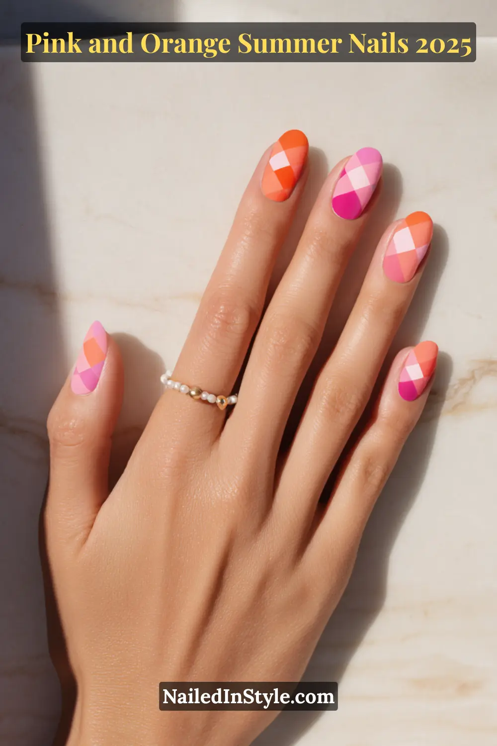 Pink and Orange Summer Nails 2026: Sunset Tips, Endless Vibes 12 Hand with almond-shaped matte nails featuring a checkerboard pattern in alternating shades of pink and orange, resting on a marble surface with a pearl-accent ring on fair skin.