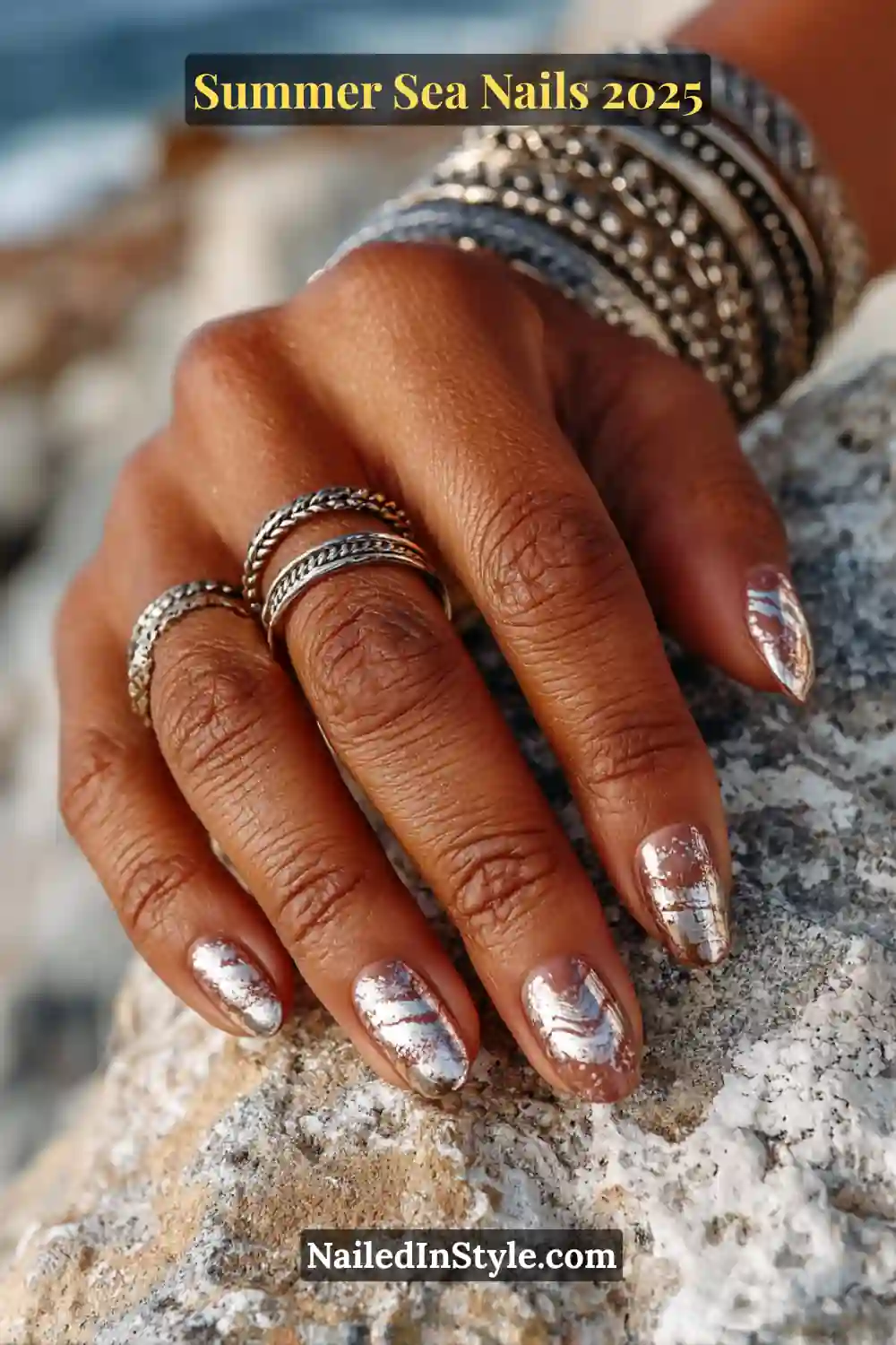 Hand with chrome wave accent nail design in metallic silver and nude, styled with silver rings on a rocky seaside background