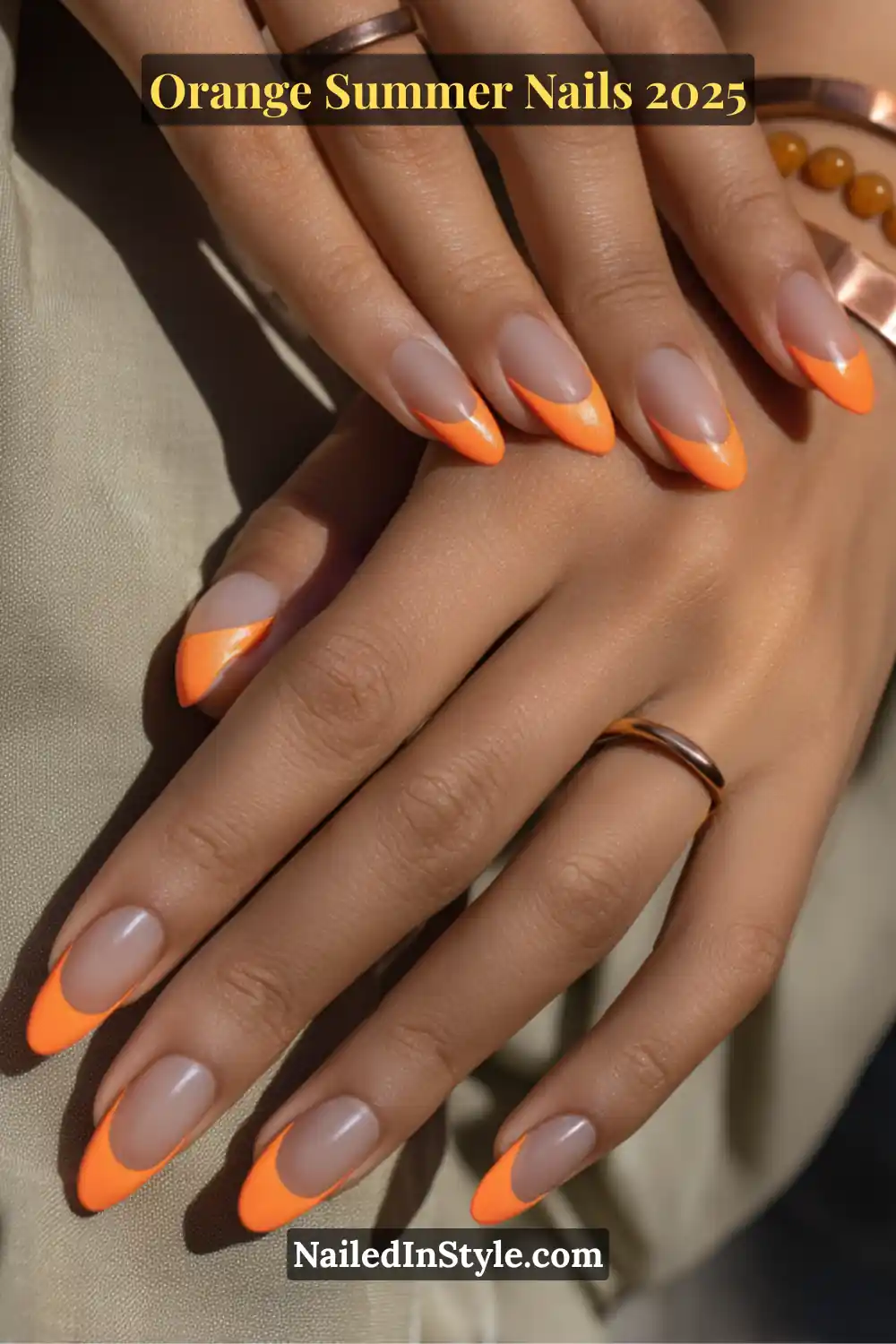 Close-up of almond-shaped nails with a soft nude base and vibrant orange French tips, styled with minimal copper rings.