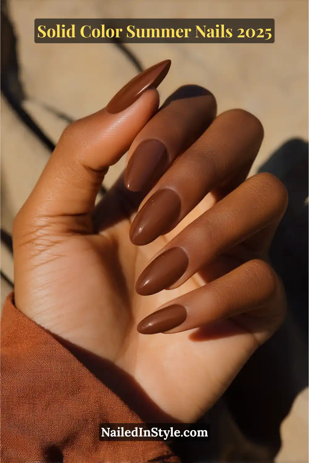 A hand with long almond-shaped nails painted in a glossy cocoa brown polish, shown against deep skin and warm-toned clothing in sunlight.