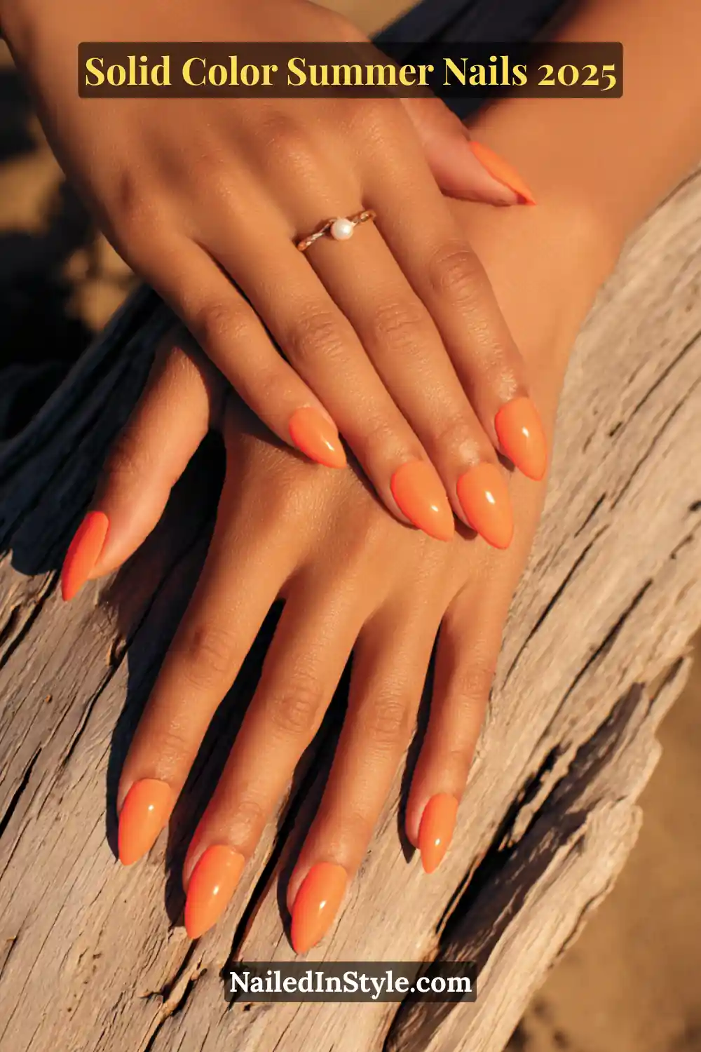 Two hands with almond-shaped nails painted in a glossy coral reef orange shade, resting on sunlit driftwood.