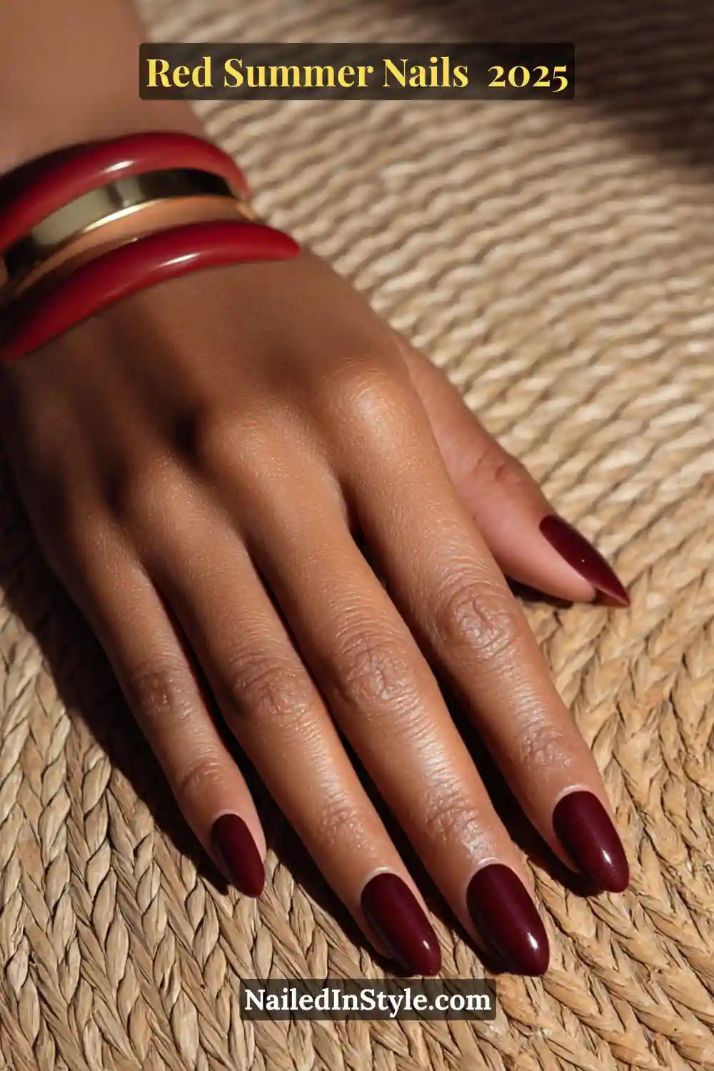 Red Summer Nails 2026: Bold, Fiery & Classic Warm-Weather Looks 9 Hand with deep crimson gel oval nails on brown skin, paired with bold red and gold bracelets on a woven mat background