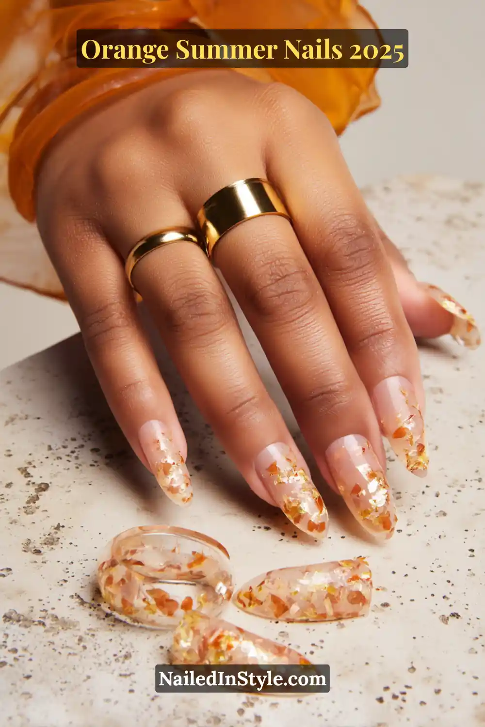 Close-up of a hand with long transparent nails decorated with metallic orange and gold foil flakes, paired with bold gold rings.