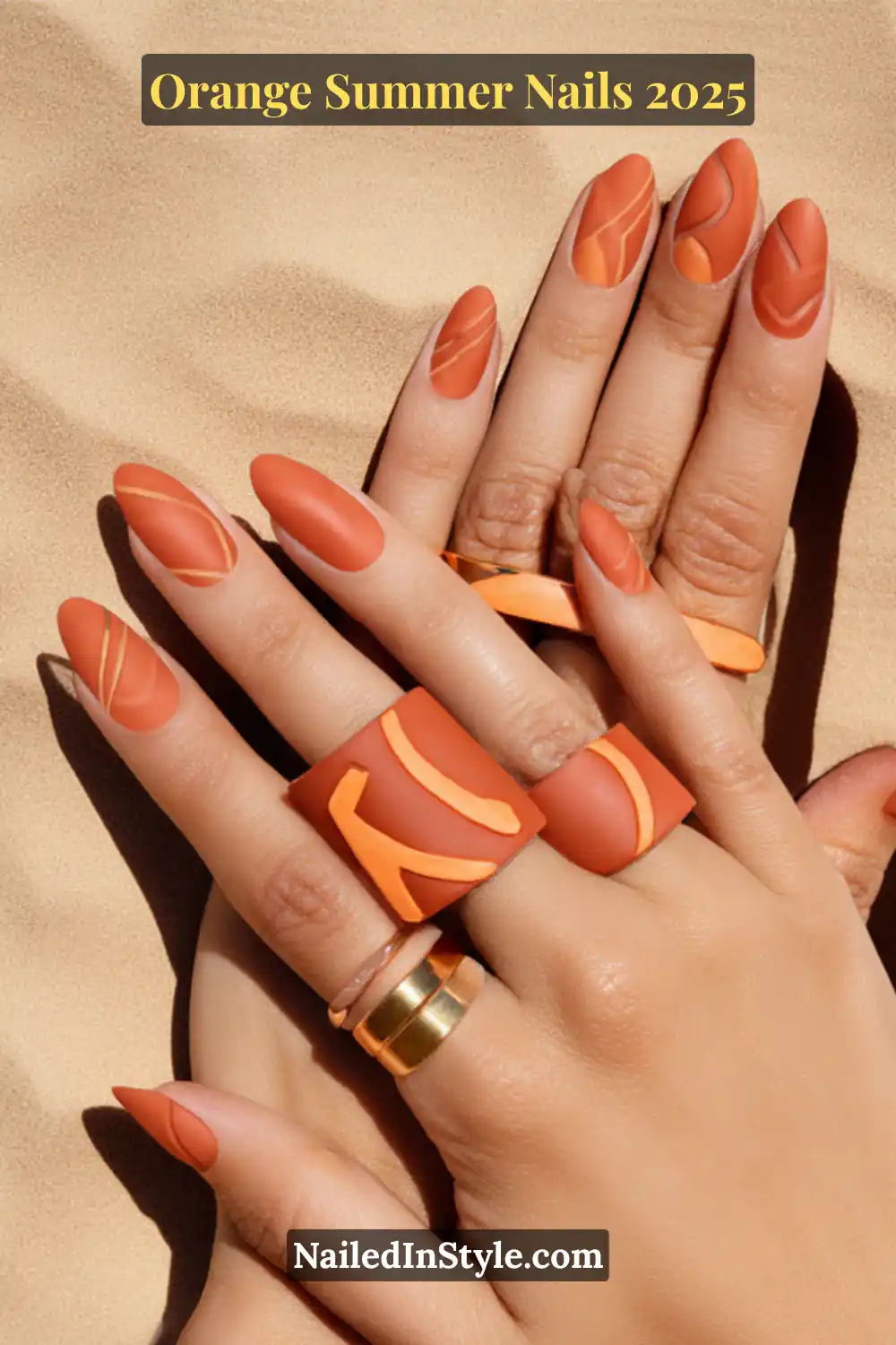 Hands with matte burnt-orange almond nails featuring minimalist orange abstract line art, styled with bold geometric rings on a sandy background.
