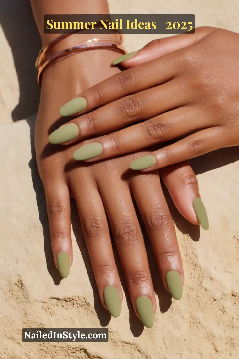 Hands with almond-shaped nails painted in soft matte olive green, resting on sandy stone.