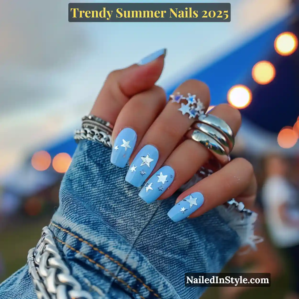Trendy Summer Nails 2026: Top Designs, Colors & Ideas for the Season 29 Close-up of a fair-toned hand with sky blue square nails decorated with silver star decals, styled with chunky silver rings and denim at an outdoor music festival.