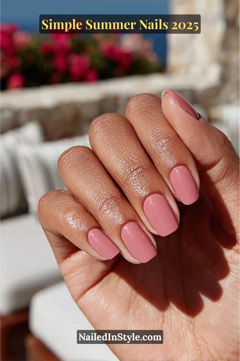 Glossy mauve-pink nails on a medium-to-deep skin tone hand, styled with a scenic stone patio and ocean view in the background.