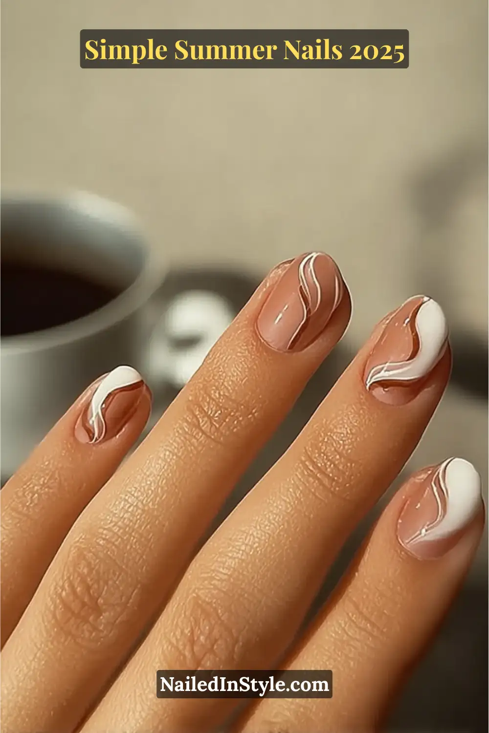 A hand with medium-toned skin displays short almond nails with a glossy nude base and flowing milky white swirl designs, set against a soft café background.