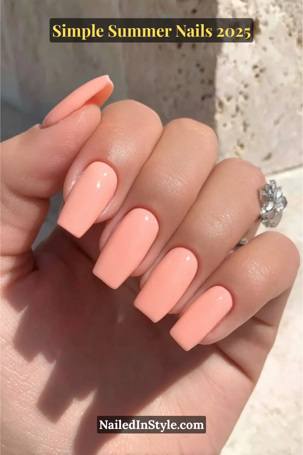 A hand with neutral-toned skin displays long square nails painted in a glossy peach polish, posed against a sunlit marble background.