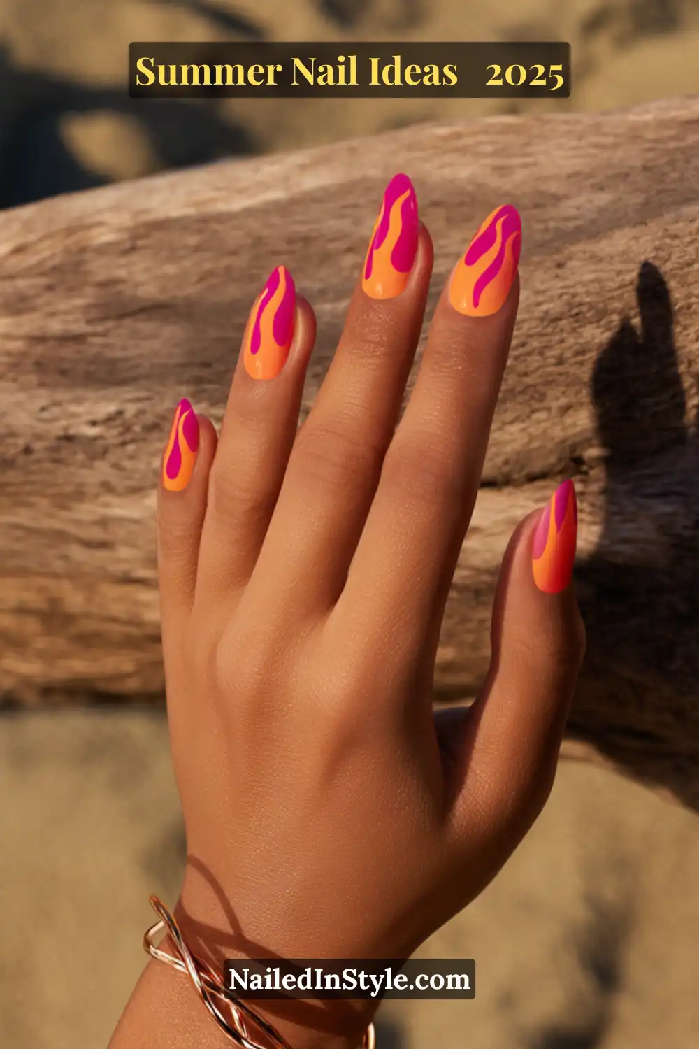 Close-up of a golden-toned hand with almond-shaped nails painted in a high-gloss orange base featuring hot pink flame designs, posed against a sunlit driftwood background on the beach.