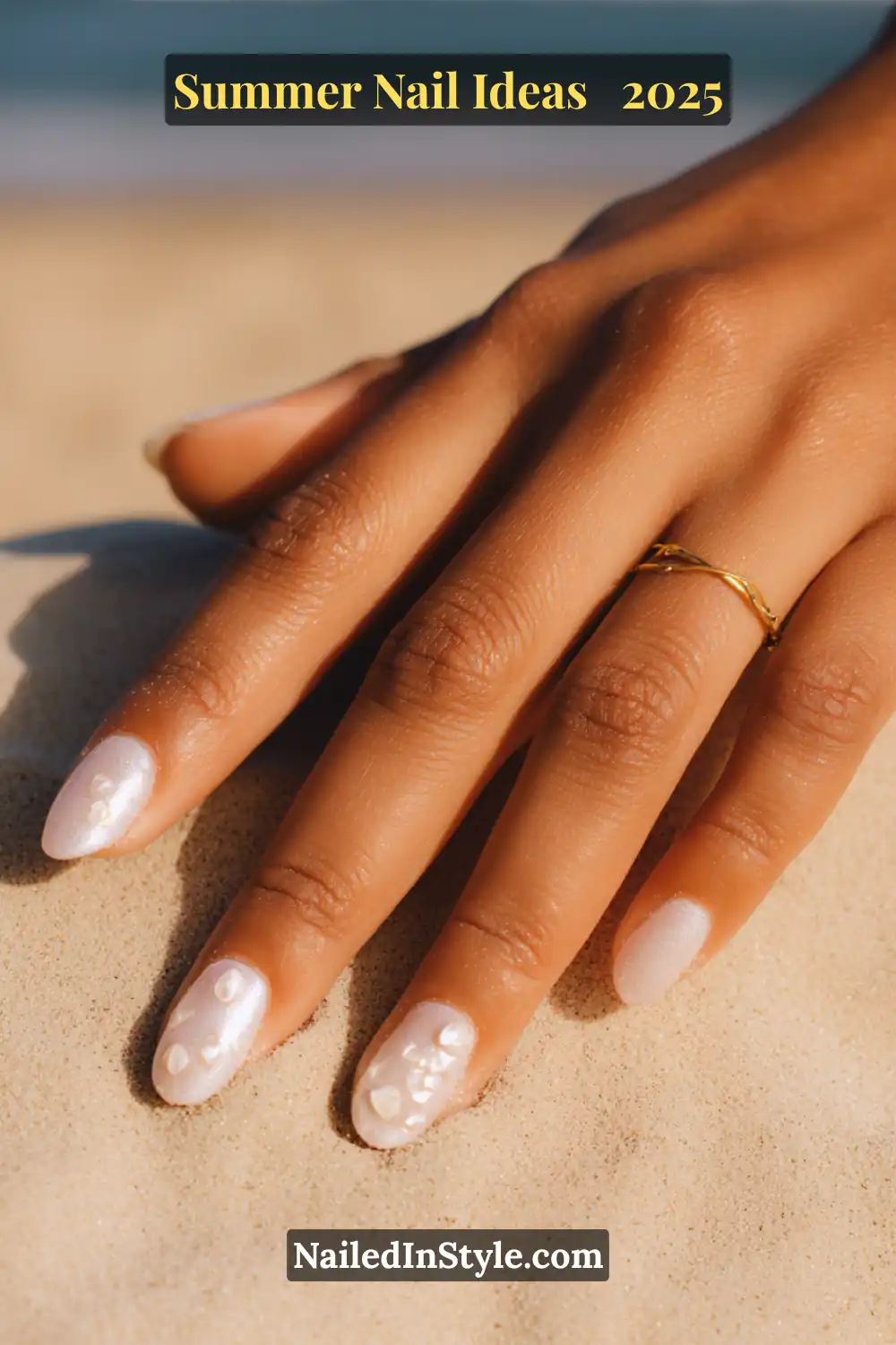 Close-up of a golden-tan hand with almond-shaped nails featuring pearlescent white chrome polish and 3D baby shell accents, resting on soft beach sand with blurred ocean in background.