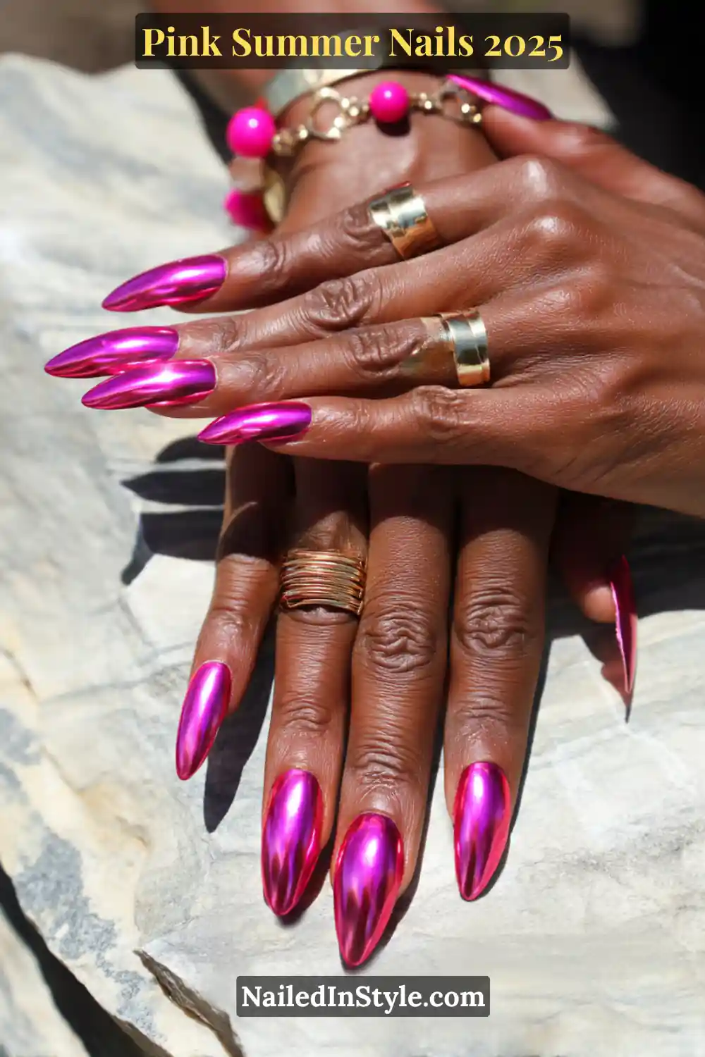 Close-up of dark skin hands with long almond-shaped chrome nails in hot pink, accessorized with bold gold rings and a pink beaded bracelet.