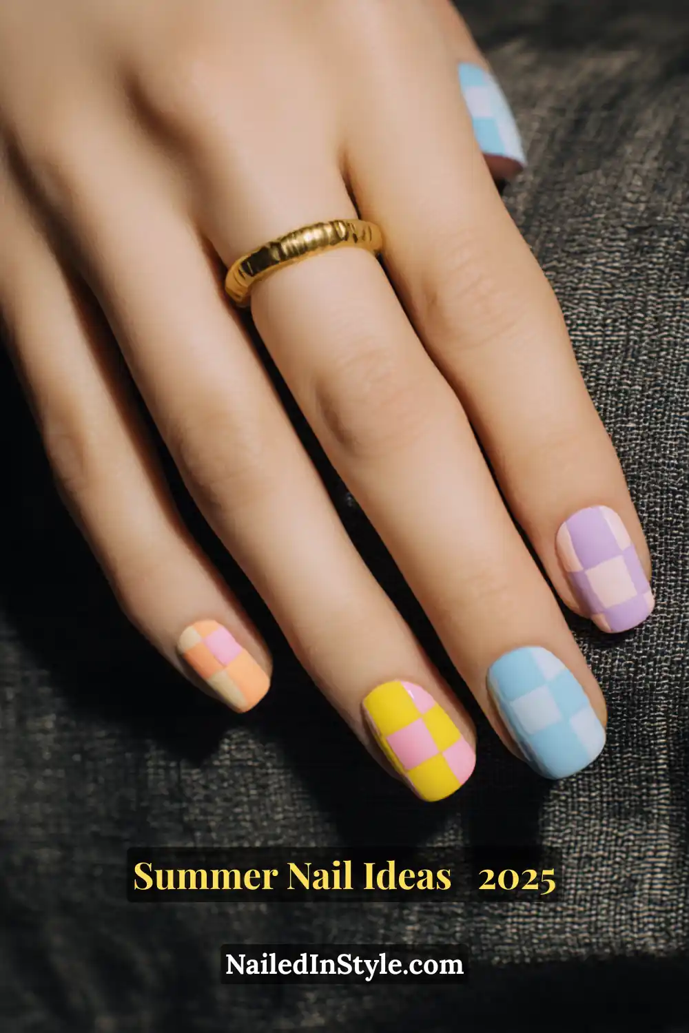 Close-up of an ivory-skinned hand with square-shaped nails featuring pastel checkerboard nail art in yellow, pink, peach, lilac, and baby blue blocks, set against a textured dark fabric background.