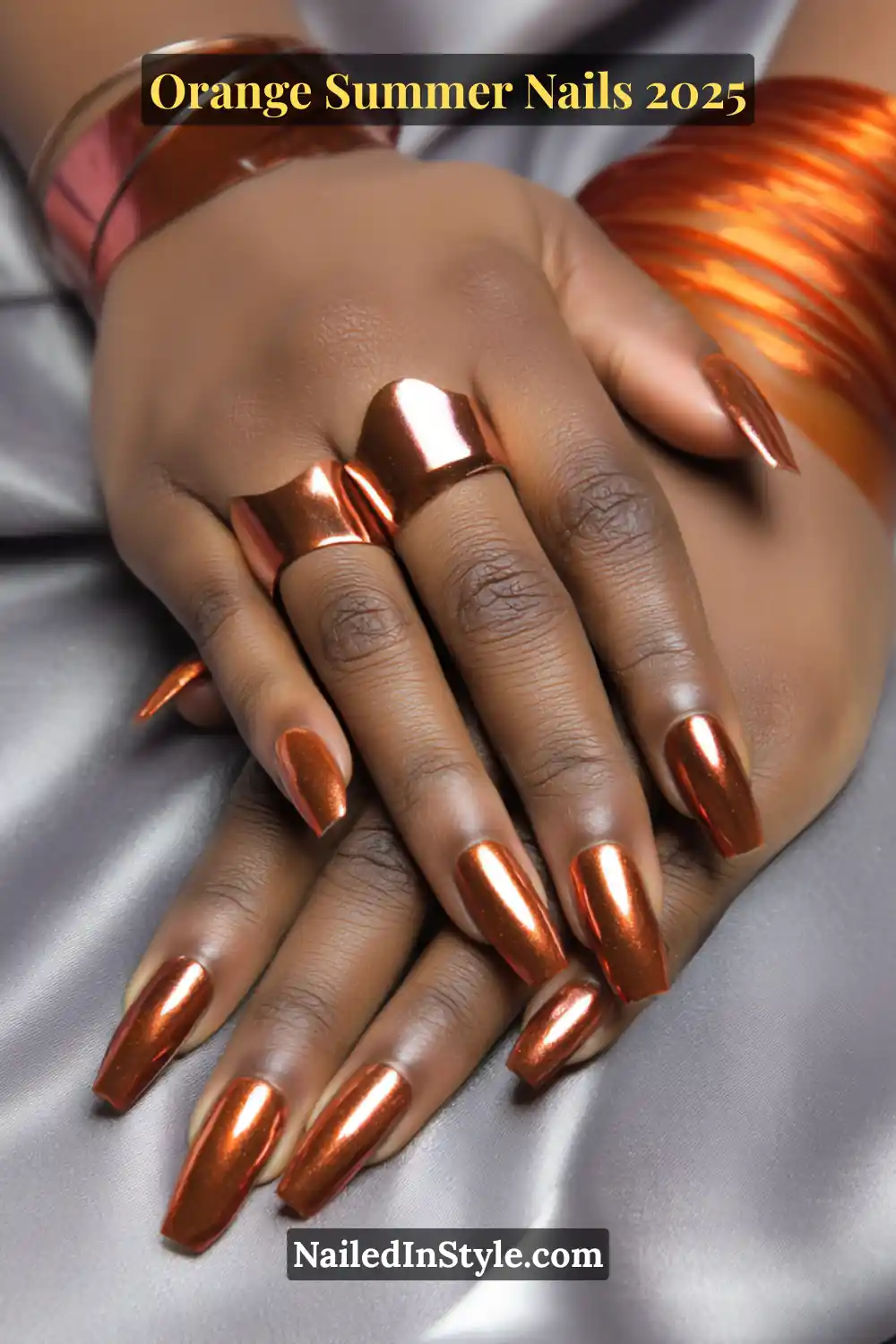 Close-up of deep skin tone hands with long chrome orange coffin nails and bold metallic rings on a satin silver backdrop.