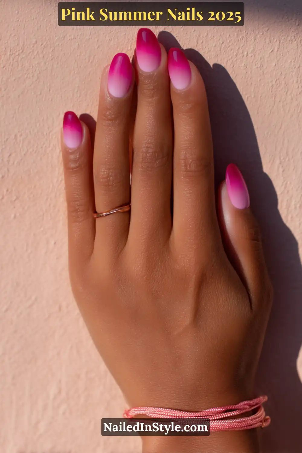 A hand with almond-shaped nails featuring a seamless magenta-to-white ombré fade, styled with a rose gold ring and pink cord bracelet.