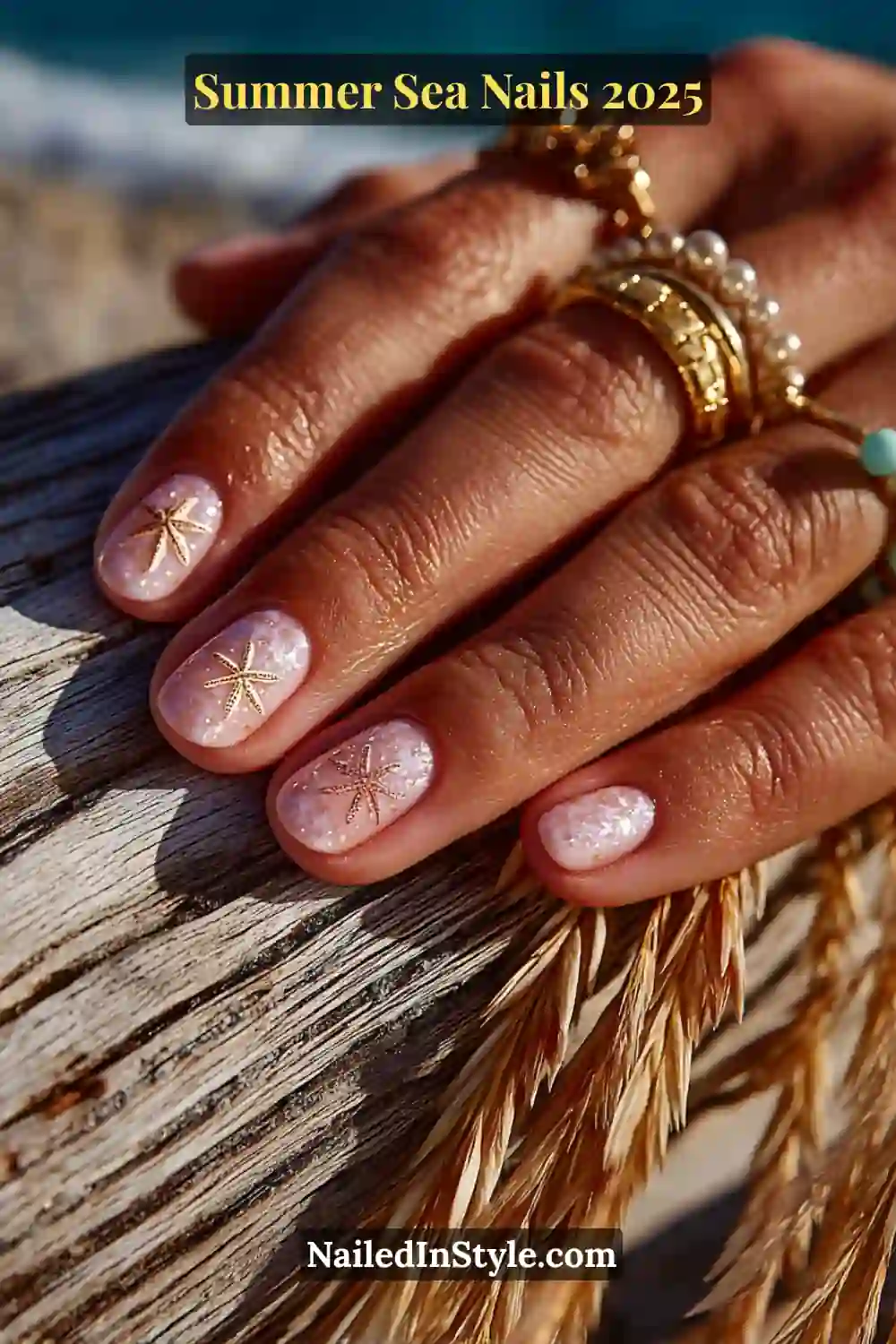 Close-up of nails with tiny gold starfish decals on seashell-pink polish, hand resting on driftwood with beach rings
