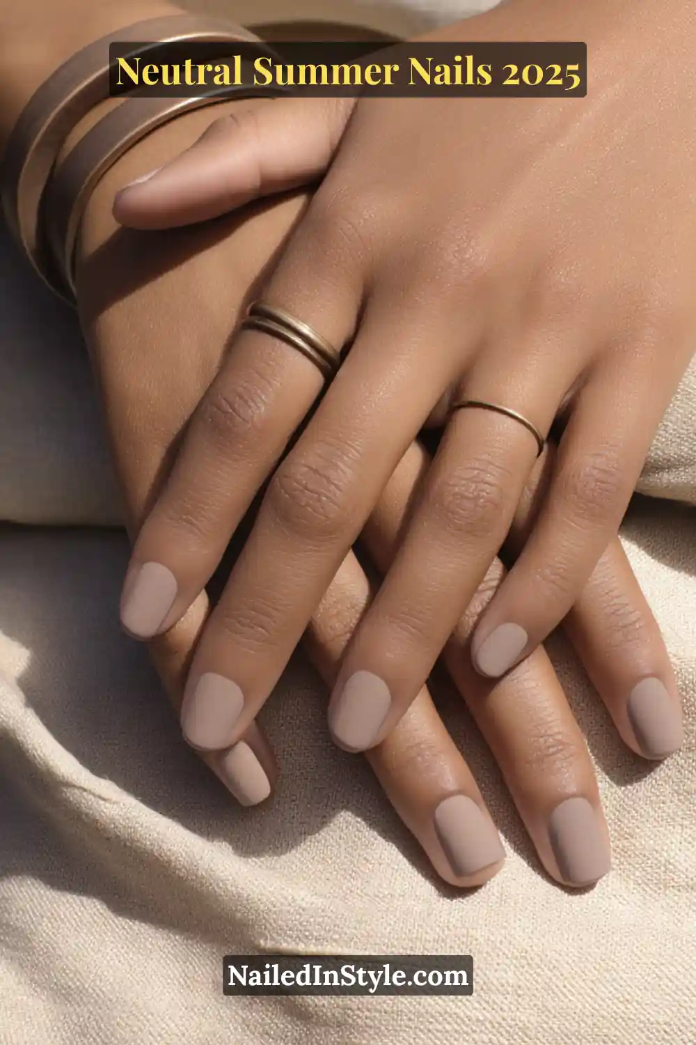 Neutral Summer Nails 2026: Clean, Chic & Warm-Weather Ready 12 Close-up of hands with matte taupe nail polish in a soft square shape, styled with bronze rings on warm tan skin, resting on a neutral fabric.