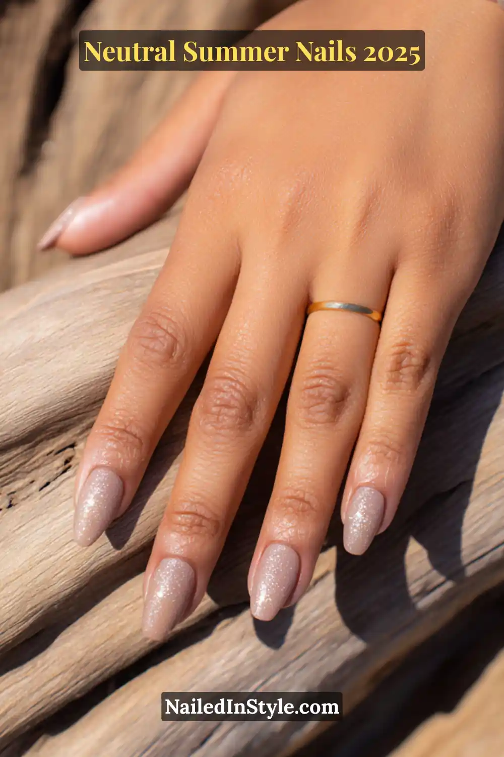 Neutral Summer Nails 2026: Clean, Chic & Warm-Weather Ready 23 Close-up of a hand with almond-shaped nails painted in matte nude polish with a fine layer of silver micro glitter, resting on driftwood.