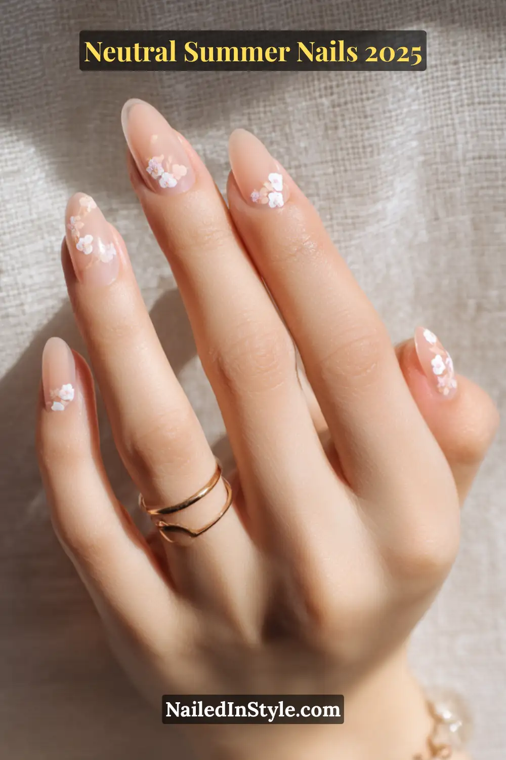 Neutral Summer Nails 2026: Clean, Chic & Warm-Weather Ready 25 Close-up of fair-toned hand with almond-shaped nude nails adorned with delicate white floral decals, styled with gold rings.