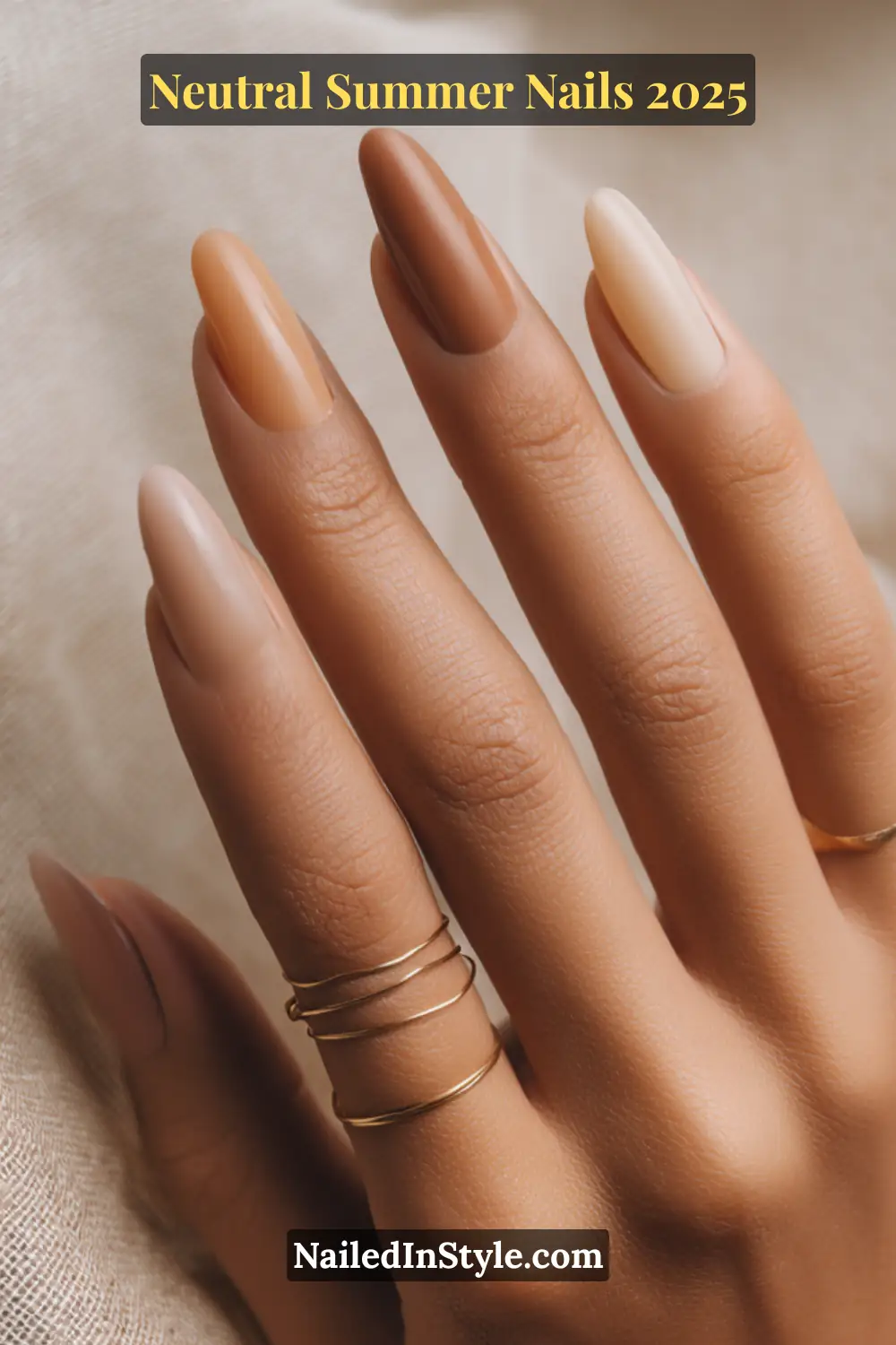 Neutral Summer Nails 2026: Clean, Chic & Warm-Weather Ready 3 Medium-toned hand showcasing almond-shaped nails in a gradient of sandy beige, caramel, and tan rose shades, styled with delicate gold rings on a neutral fabric background.