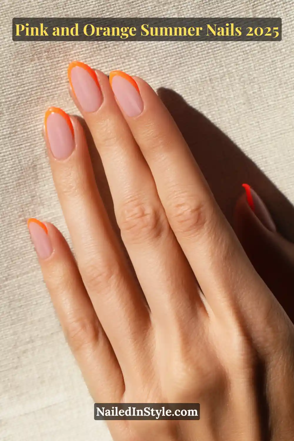 Pink and Orange Summer Nails 2026: Sunset Tips, Endless Vibes 8 Hand with almond-shaped nails featuring a sheer baby pink base and bright orange French tips, shown side angle on fair skin against sunlit linen fabric.