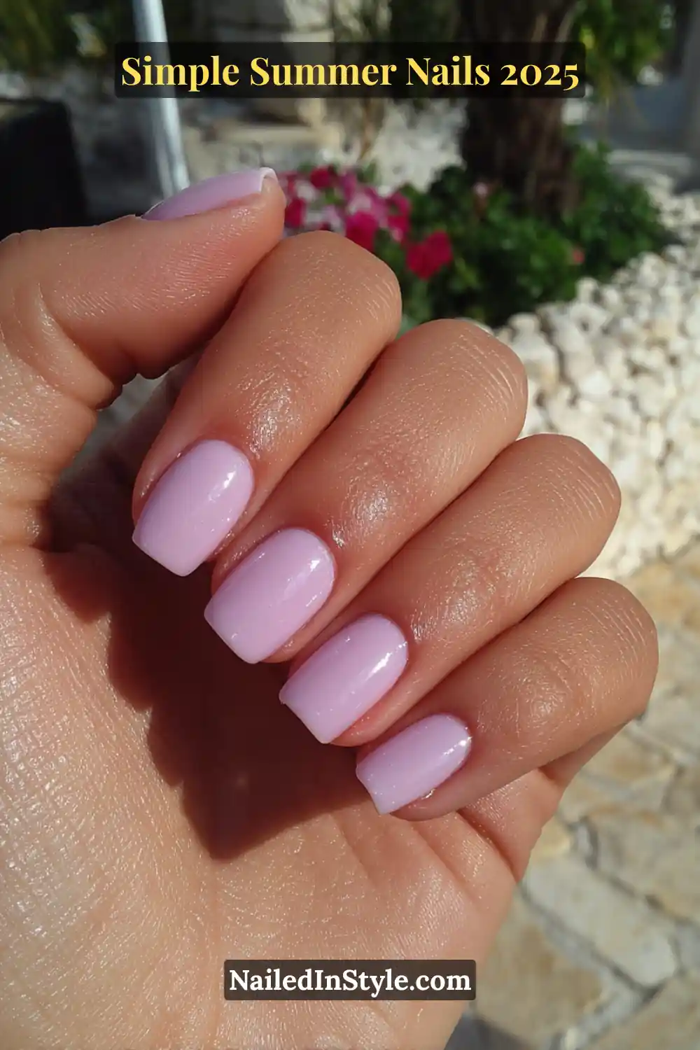Close-up of a hand with pale lilac square-shaped nails, photographed in bright sunlight with a natural stone patio and flowers in the background.
