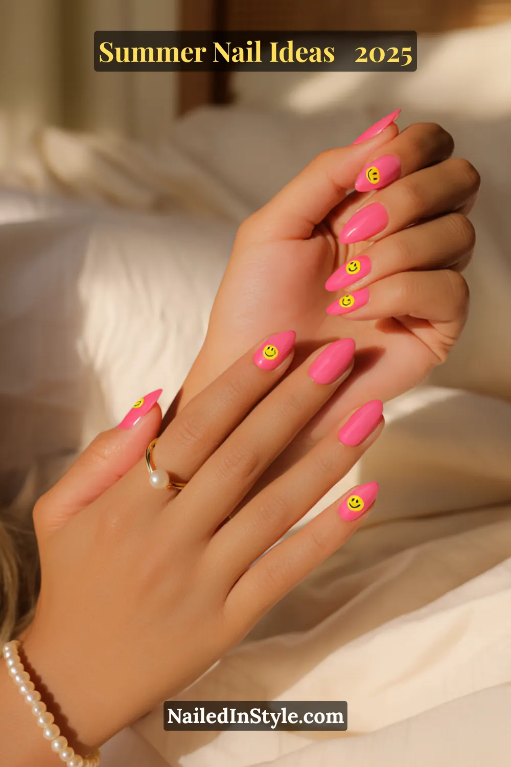 Close-up of fair peach-toned hands with almond-shaped nails painted in glossy candy pink polish, each featuring a bold yellow smiley face decal. Background includes soft beige bedding and warm morning sunlight.