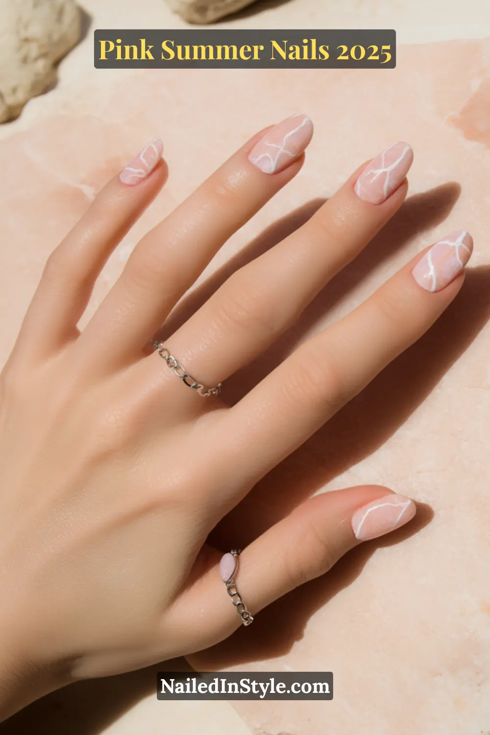 Close-up of short nails with soft pink marble design, styled with silver rings on a neutral stone background.