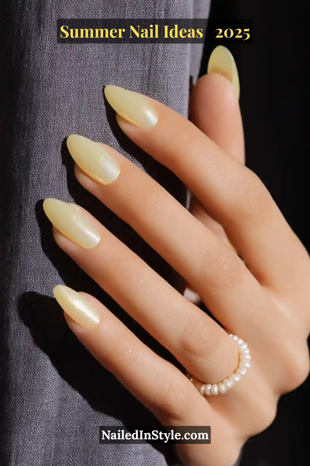 Close-up of a porcelain-toned hand with almond-shaped nails in a sheer lemonade-yellow jelly polish with a luminous chrome finish, styled against a dark linen backdrop with a pearl ring.