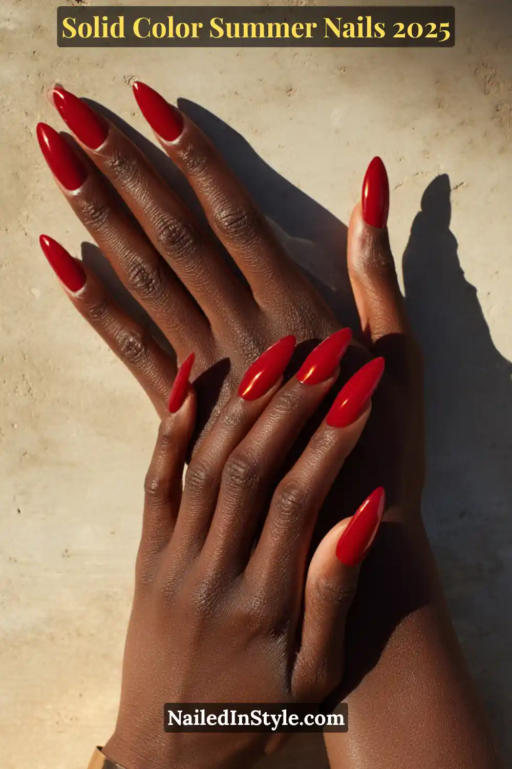 Two hands with long almond-shaped nails painted in a high-shine, deep red lacquer, shown against sunlit dark skin on a neutral stone background.