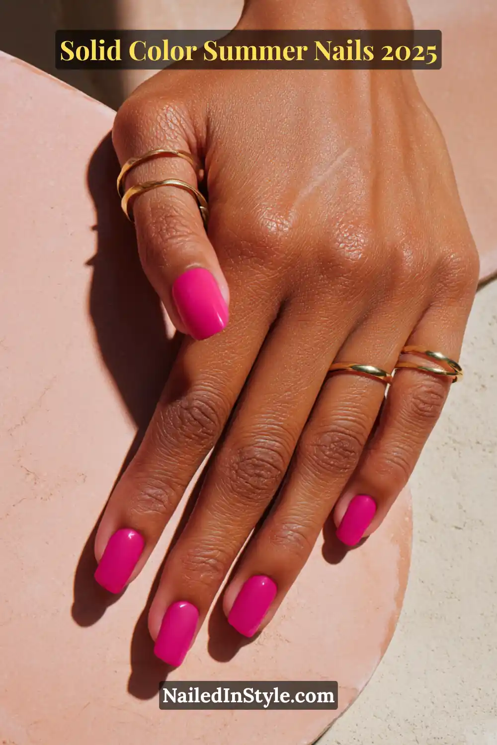 A single hand with short square nails painted in a glossy, vibrant fuchsia pink gel polish, accessorized with gold rings, resting on a pink tile.