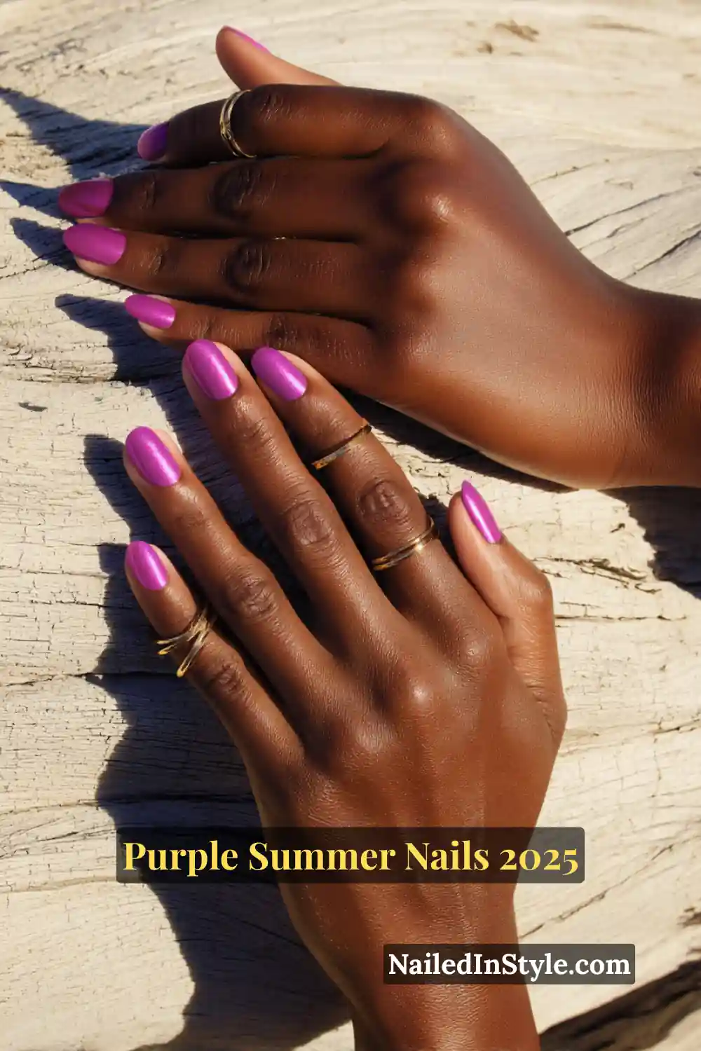 A pair of hands with deep skin tone featuring vibrant fuchsia-purple nail polish, resting on a sunlit piece of driftwood.