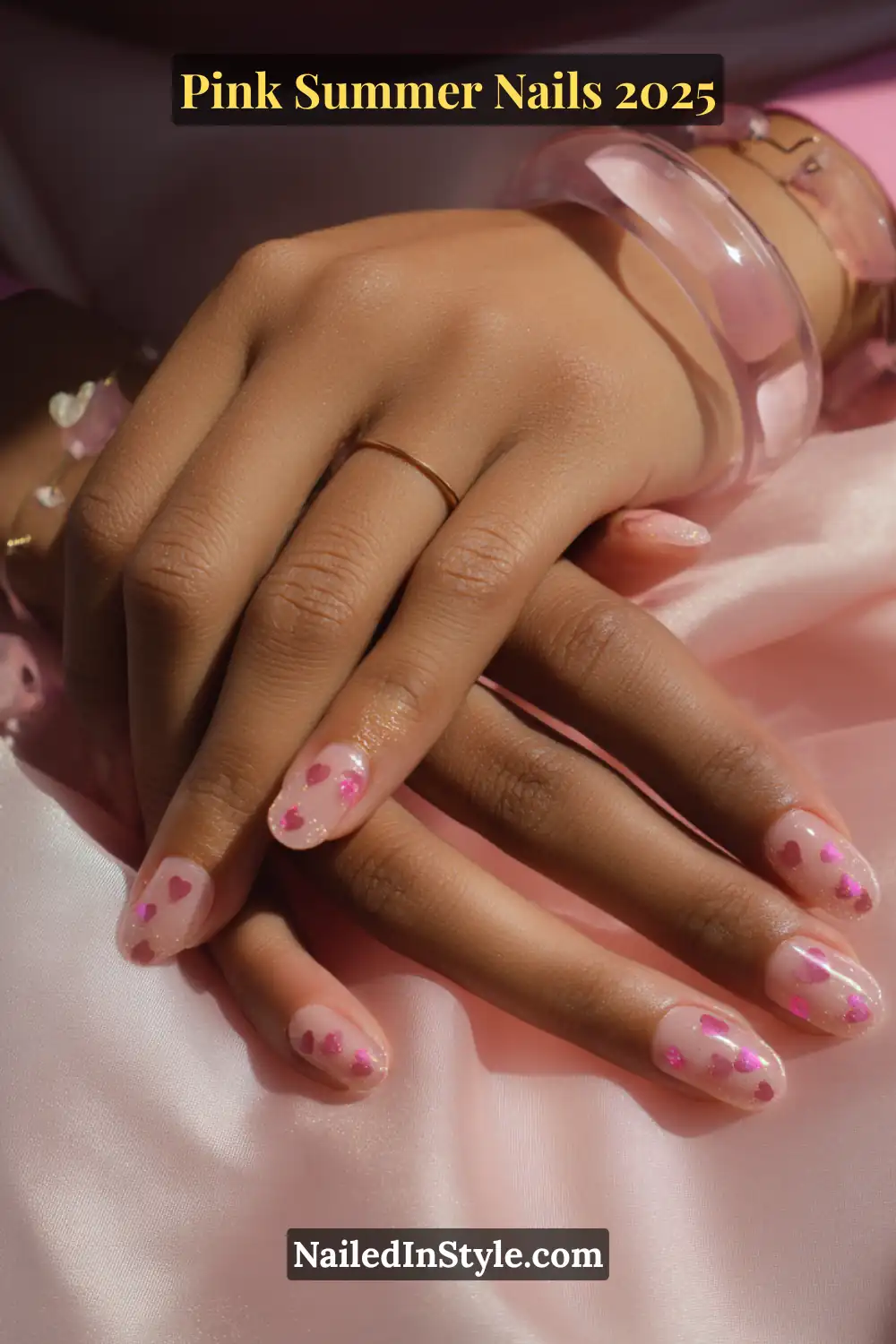 Close-up of a hand with almond-shaped nails featuring translucent nude polish and scattered pink glitter hearts, styled with soft pink and gold accessories.