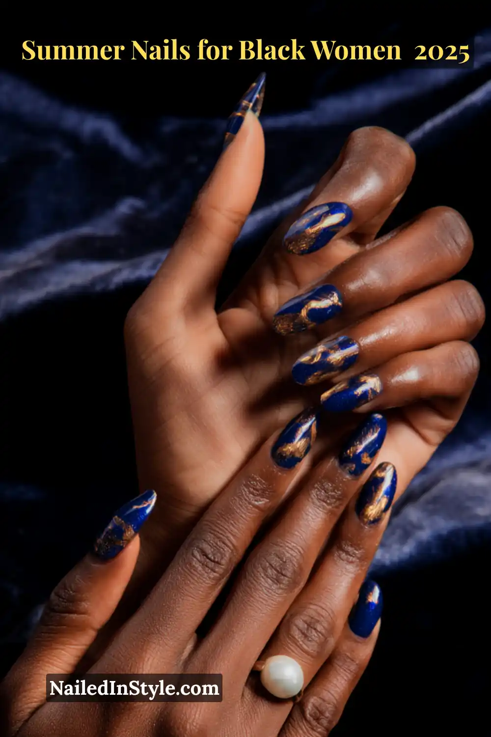 Close-up of a Black woman's hands featuring oval cobalt blue nails with abstract gold foil accents, paired with a pearl ring on a velvet background.