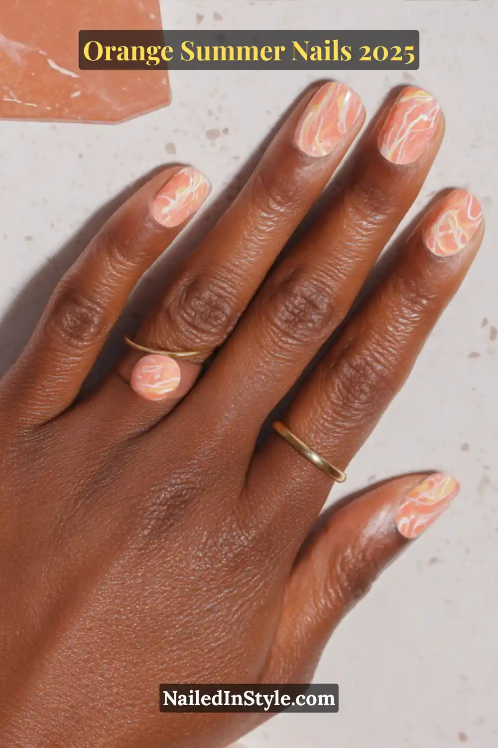 Close-up of short natural nails with orange and white marble swirl nail art, styled with minimalist gold rings on deep skin tone.