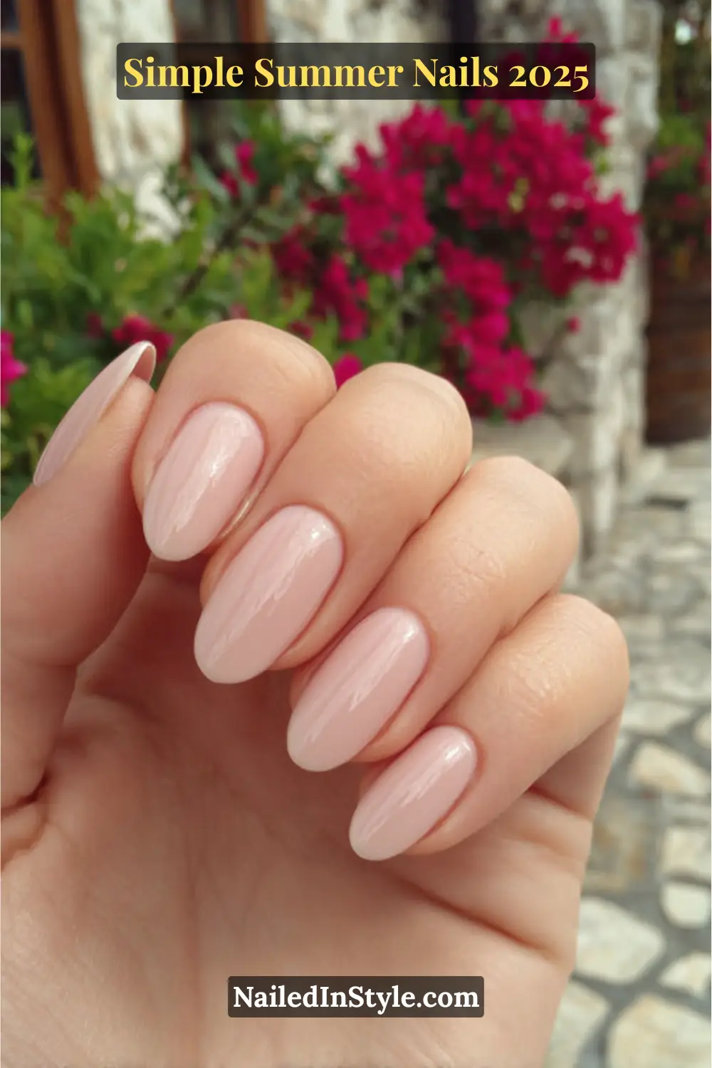 Hand with almond-shaped nude pink nails, glossy finish, held outdoors near stone wall and vibrant pink flowers.
