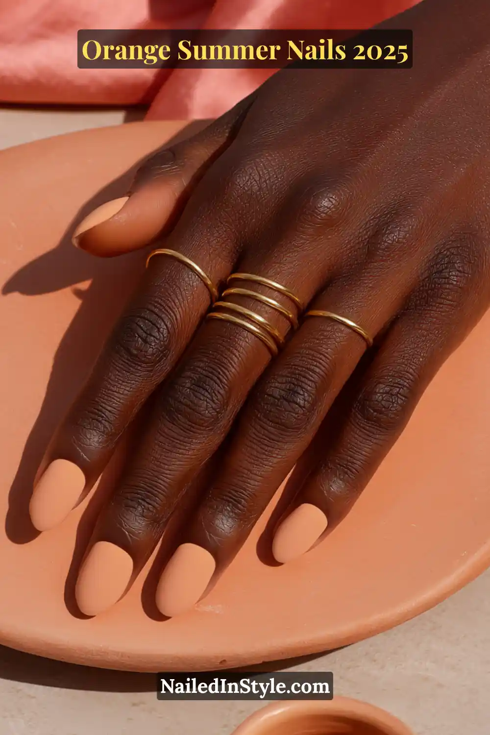 Close-up of short, rounded nails in a creamy matte orange shade on dark skin, accented with minimal gold rings on terracotta background.