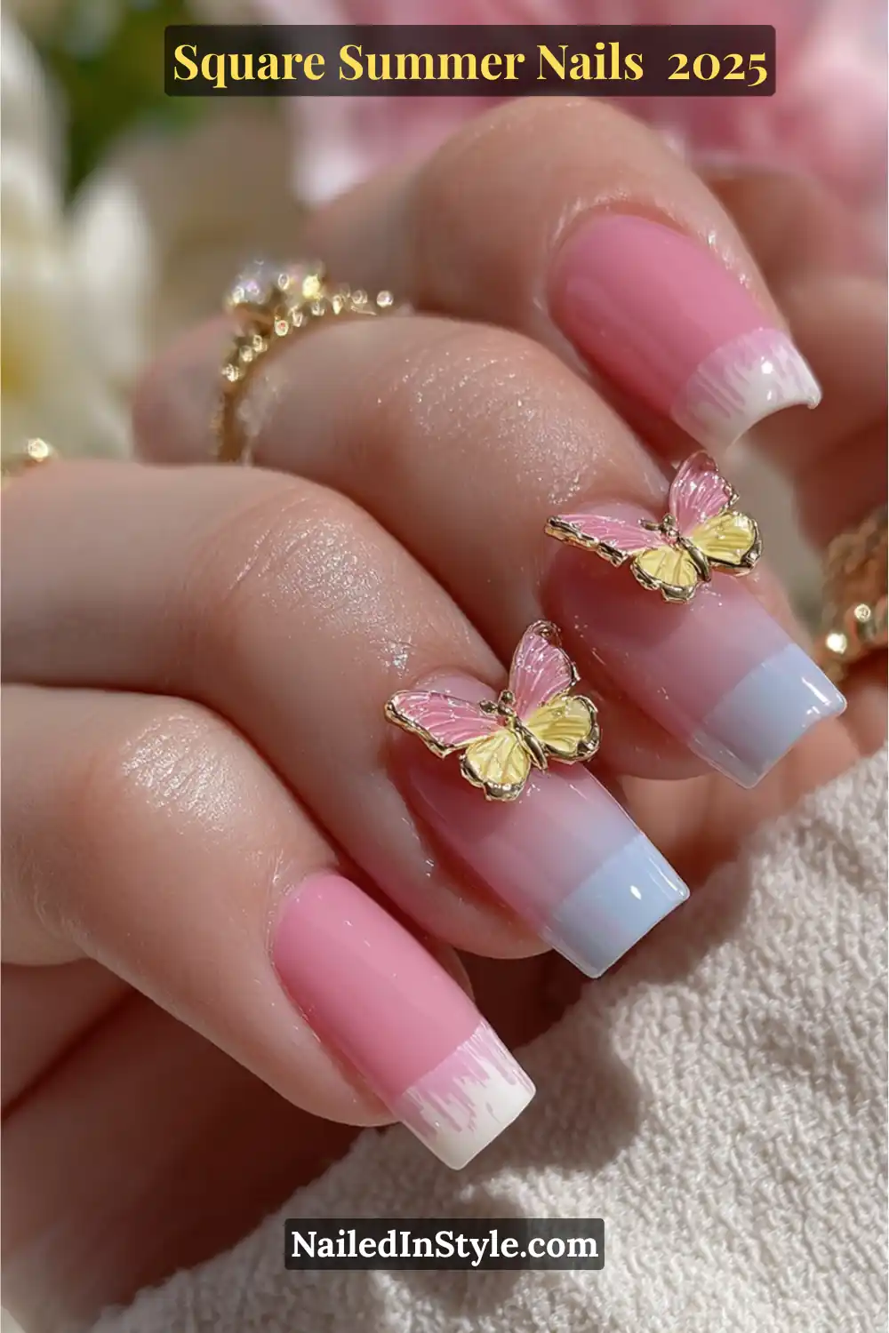 Square pink nails with pastel blue ombré tips and gold-accent butterfly charms on a woman’s hand in natural sunlight