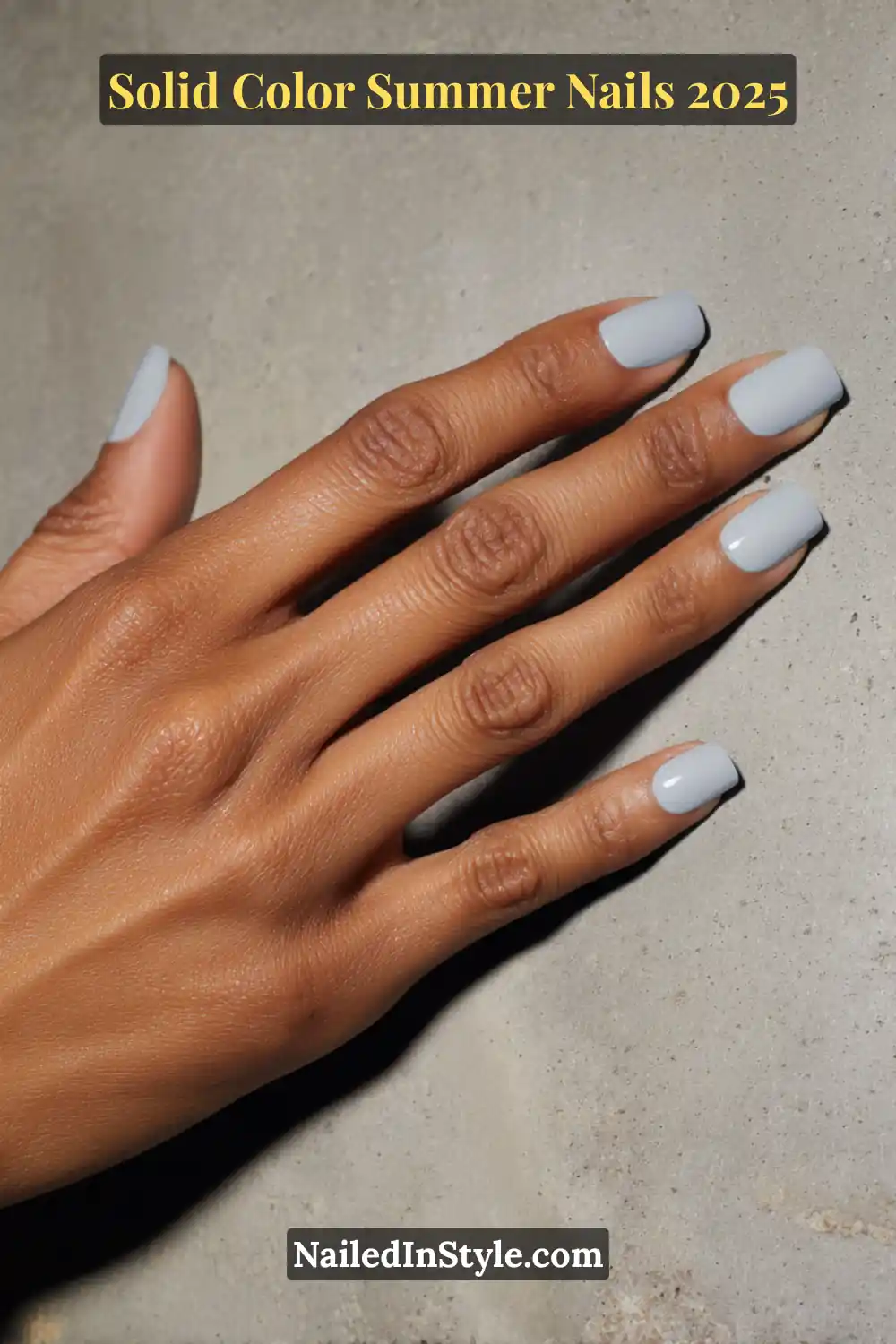 Hand with short square nails painted in a soft stone grey gel polish against a smooth cement background.