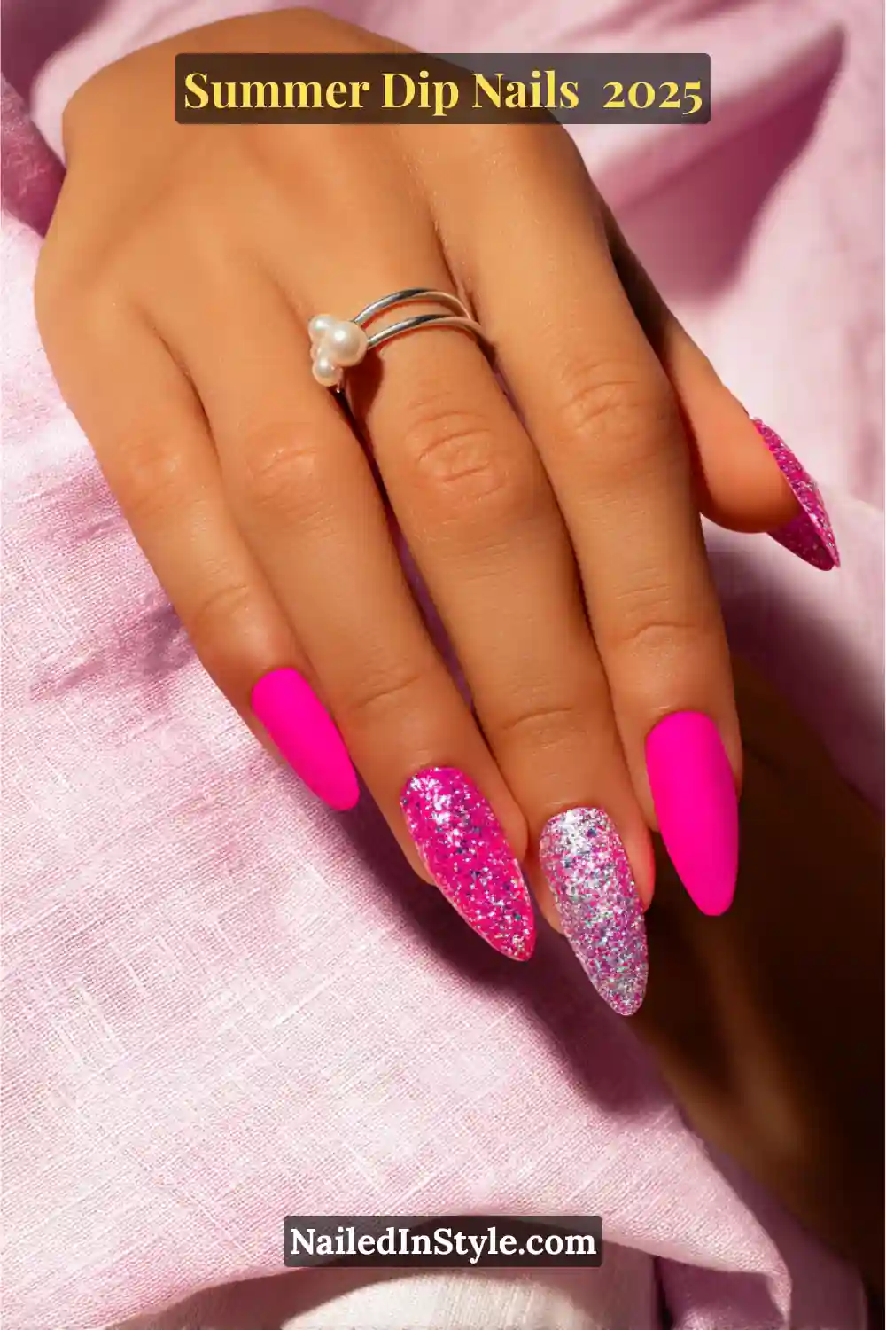 Long stiletto dip nails in hot pink with accent nails in pink and silver glitter, styled against soft pink fabric.