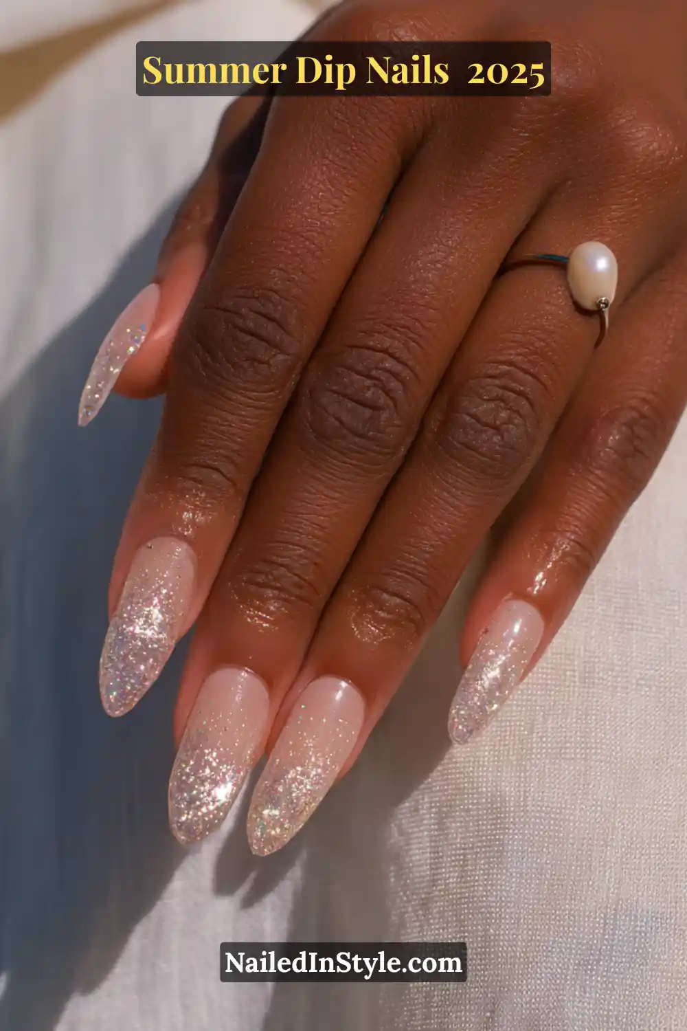 Almond-shaped nails with a soft clear base and silver glitter-dipped tips on deep skin, styled with a pearl ring.