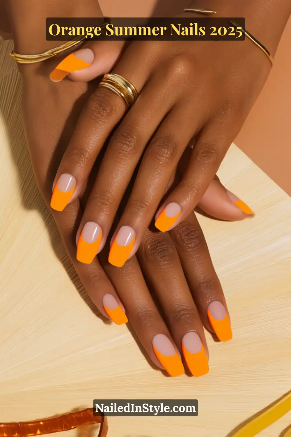 Close-up of hands with square-tipped nails featuring bold neon-orange polish and half-moon cuticle designs on a nude base, paired with gold rings.