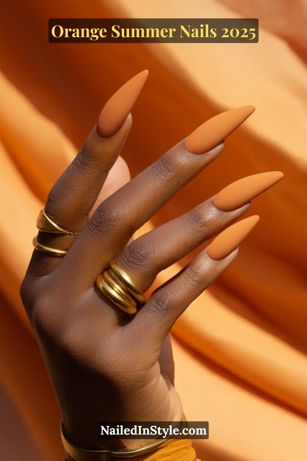 Elegant hand with deep skin tone showcasing matte mango orange coffin nails and stacked gold rings on a flowing orange fabric background.